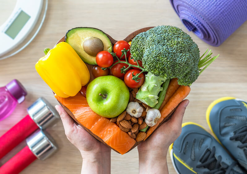 heart shaped cutting board with fruits, vegetables, nuts and salmon with athletic shoes and hand weights in background