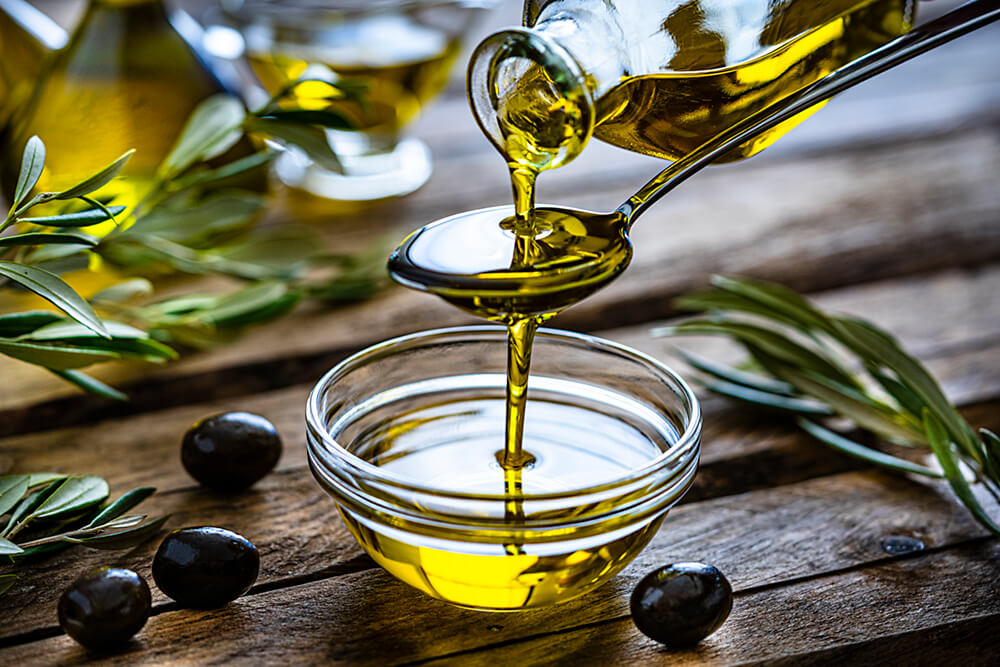 Photo of olive oil pouring into spoon over glass bowl on wooden table with black olvies
